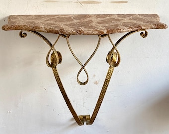 Puede incluir: Una consola de pared con una superficie de mármol pulido en tonos marrón y blanco con un borde suave y curvado. La estructura de soporte está hecha de metal dorado martillado, con volutas intrincadas y un marco en forma de V.