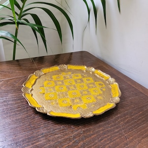 Op de afbeelding: Een ronde, gouden dienblad met een gele rand en een ingewikkeld, gegraveerd ontwerp. Het dienblad ligt op een houten oppervlak.