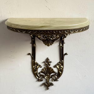 May include: A decorative wall shelf with a half-moon, light-coloured marble top. The shelf is supported by ornate, bronze-coloured metalwork with intricate scroll and floral designs. The shelf is mounted on a white wall.