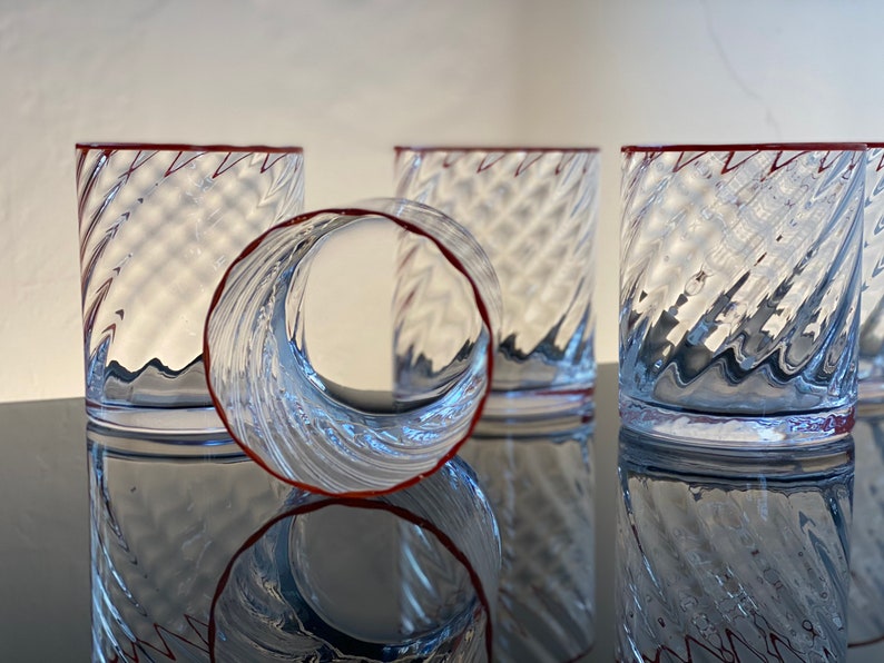 May include: Four clear glass tumblers with a red rim and a swirling, textured design. The tumblers are stacked on top of each other.