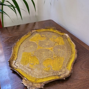May include: A decorative, antique serving tray with a yellow and gold color scheme. The tray features an ornate, scalloped edge and intricate, embossed floral patterns. The tray is made of wood and has a distressed finish, giving it a vintage look.