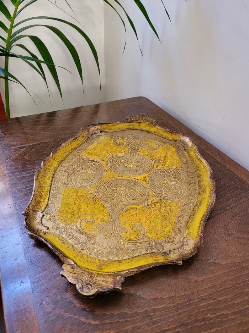 May include: A decorative, antique serving tray with a yellow and gold color scheme. The tray features an ornate, scalloped edge and intricate, embossed floral patterns. The tray is made of wood and has a distressed finish, giving it a vintage look.