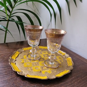 Op de afbeelding: Twee gouden gerande glazen shotglazen op een decoratief dienblad in geel en goud. Het dienblad heeft een gegolfde rand en een bloemenpatroon.