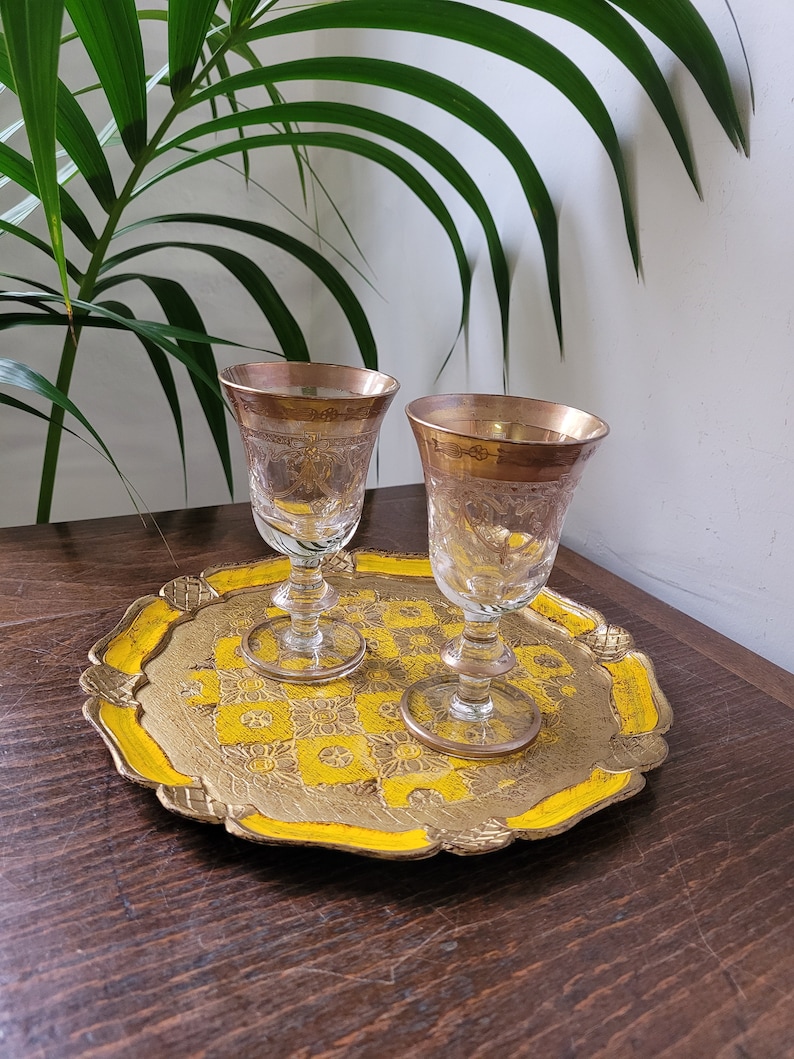 Op de afbeelding: Twee gouden gerande glazen shotglazen op een decoratief dienblad in geel en goud. Het dienblad heeft een gegolfde rand en een bloemenpatroon.