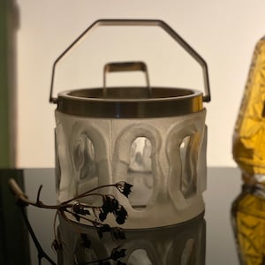 May include: A frosted glass ice bucket with a silver metal handle and rim. The bucket has a decorative design of arches around the sides.