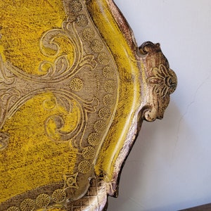May include: A decorative, ornate tray with a yellow and gold finish. The tray features intricate floral and scrollwork designs, with a distressed, textured appearance. The edges are trimmed with a darker brown, creating a vintage aesthetic.