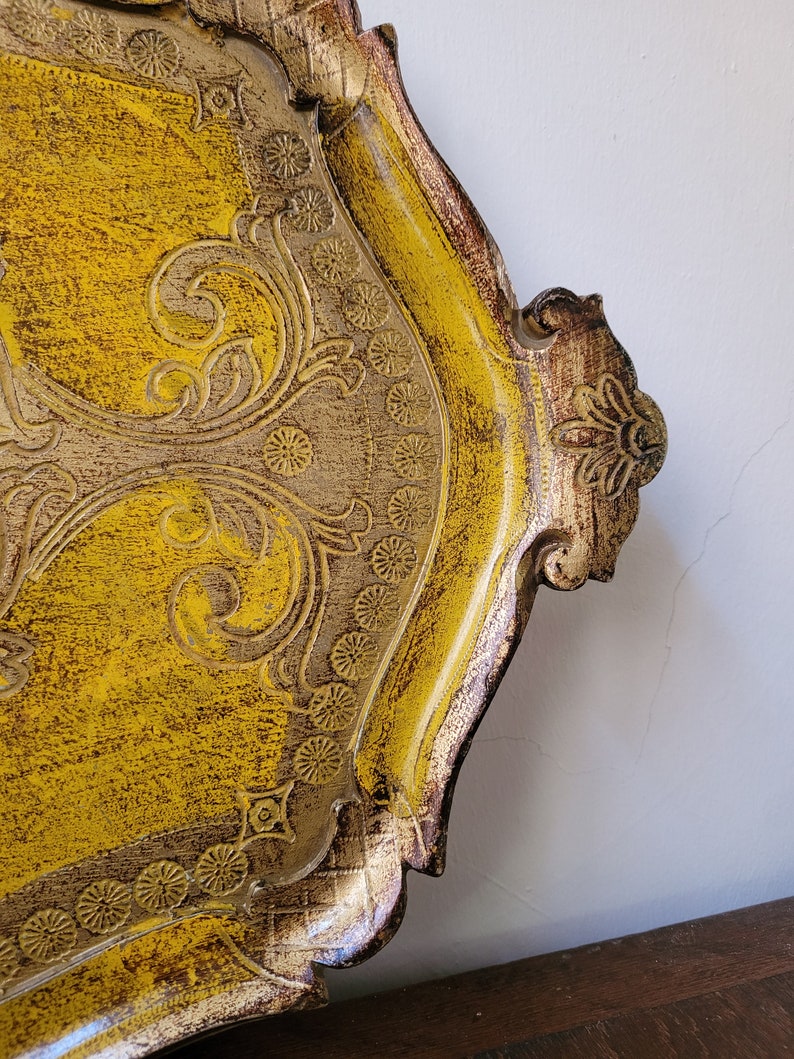 May include: A decorative, ornate tray with a yellow and gold finish. The tray features intricate floral and scrollwork designs, with a distressed, textured appearance. The edges are trimmed with a darker brown, creating a vintage aesthetic.