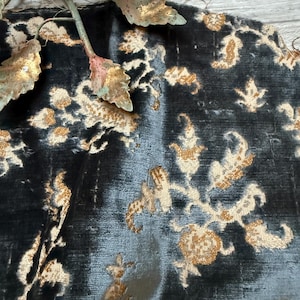 May include: Close-up of a section of dark blue fabric with a gold floral pattern. The fabric has a velvet-like texture. A decorative metal floral element with clear crystal accents is in the upper left corner.