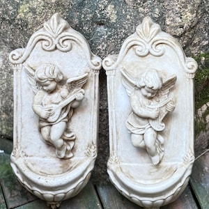 May include: Two white plaster wall plaques featuring cherubs playing musical instruments. Each plaque has a decorative frame and a scalloped bottom edge.