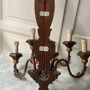 May include: A wooden chandelier with five arms and gold accents. The chandelier is wired with red and black wires and has a green and blue wire connector. The chandelier is mounted on a wall.