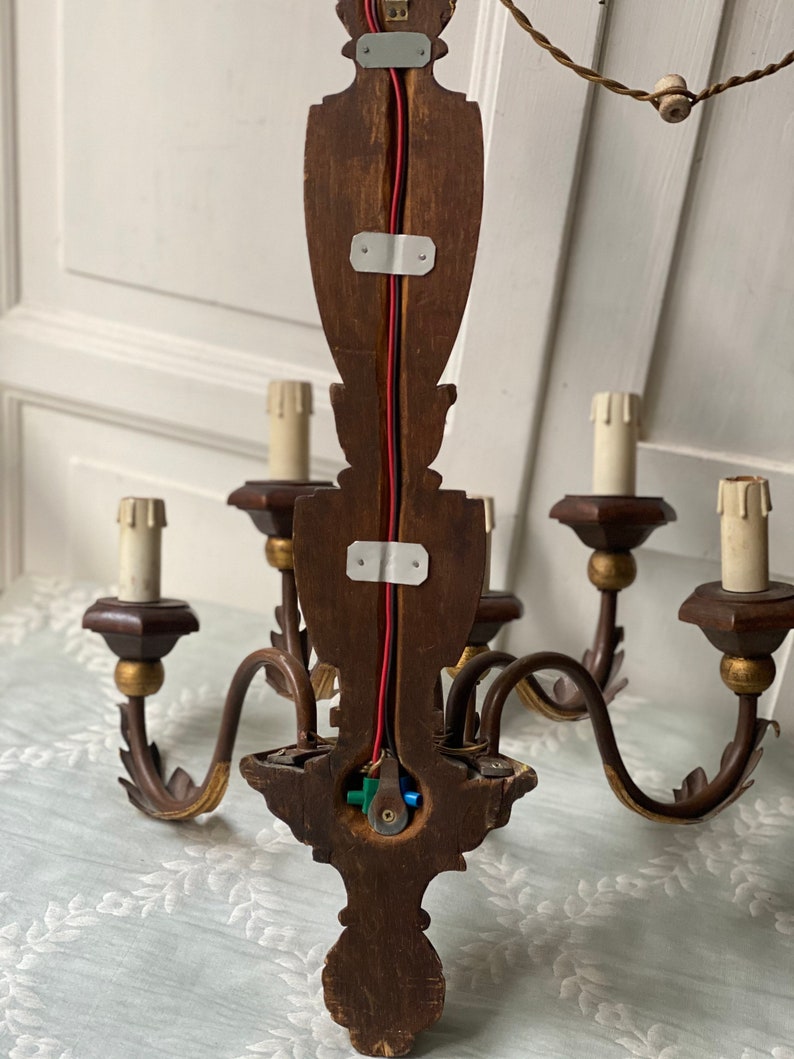 May include: A wooden chandelier with five arms and gold accents. The chandelier is wired with red and black wires and has a green and blue wire connector. The chandelier is mounted on a wall.