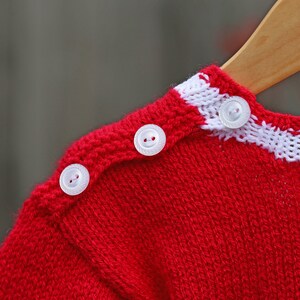 May include: A bright red knitted sweater featuring a white horizontal stripe along the neckline and shoulder. Three small, round, translucent white buttons are aligned along the shoulder seam, securing the knitted fabric in place.