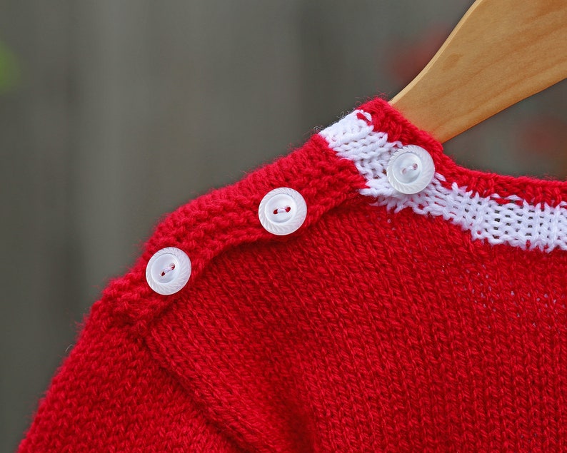 May include: A bright red knitted sweater featuring a white horizontal stripe along the neckline and shoulder. Three small, round, translucent white buttons are aligned along the shoulder seam, securing the knitted fabric in place.