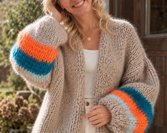 Könnte beinhalten: Eine grob gestrickte beigefarbene Strickjacke mit langen, weiten Ärmeln, die mit horizontalen Streifen in leuchtendem Orange, Türkis und Hellblau versehen sind. Das offen zu tragende Kleidungsstück besteht aus dickem Garn.