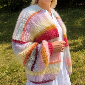Könnte beinhalten: Eine flauschige Strickjacke mit offener Vorderseite und horizontalen Streifen in Rosa, Gelb, Orange und Helllila. Das Strickkleidungsstück hat lange, lockere Ärmel und eine weiche, strukturierte Optik und wird über einem weißen Hemd getragen.