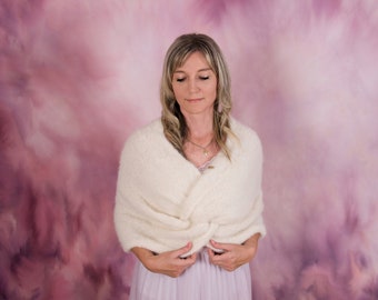 May include: A woman wearing a white knitted shawl with a knotted front. The shawl is draped over her shoulders and tied at the front. She is standing in front of a pink and purple background.