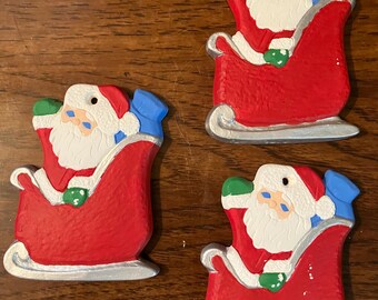 May include: Three Christmas ornaments featuring Santa Claus in a red sleigh. Each ornament has a white-bearded Santa with a red hat and coat, green mittens, and a blue sack. The sleighs have silver runners.