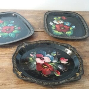 Puede incluir: Tres bandejas de metal negro con ribetes dorados y diseños florales pintados. Las bandejas presentan flores coloridas con acentos rojos, blancos, azules y verdes.