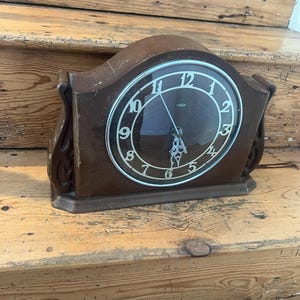 May include: A vintage brown mantel clock with a black clock face and white numerals. The clock has a curved top and decorative side panels. The clock face is protected by a glass cover. The clock is sitting on a wooden surface.