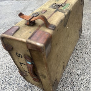 May include: A vintage rectangular suitcase in a muted olive-green fabric with worn brown leather trim, metal corner reinforcements, and a top handle. The side features the letters S.L.Z. in black, with metal latches and hinges along the top edge.
