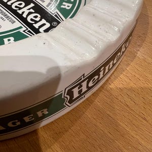May include: White ceramic ashtray featuring the Heineken logo in green and black. The ashtray is circular with a raised edge and a grooved design. The words "LAGER BEER" and "Heineken" are displayed on the surface.