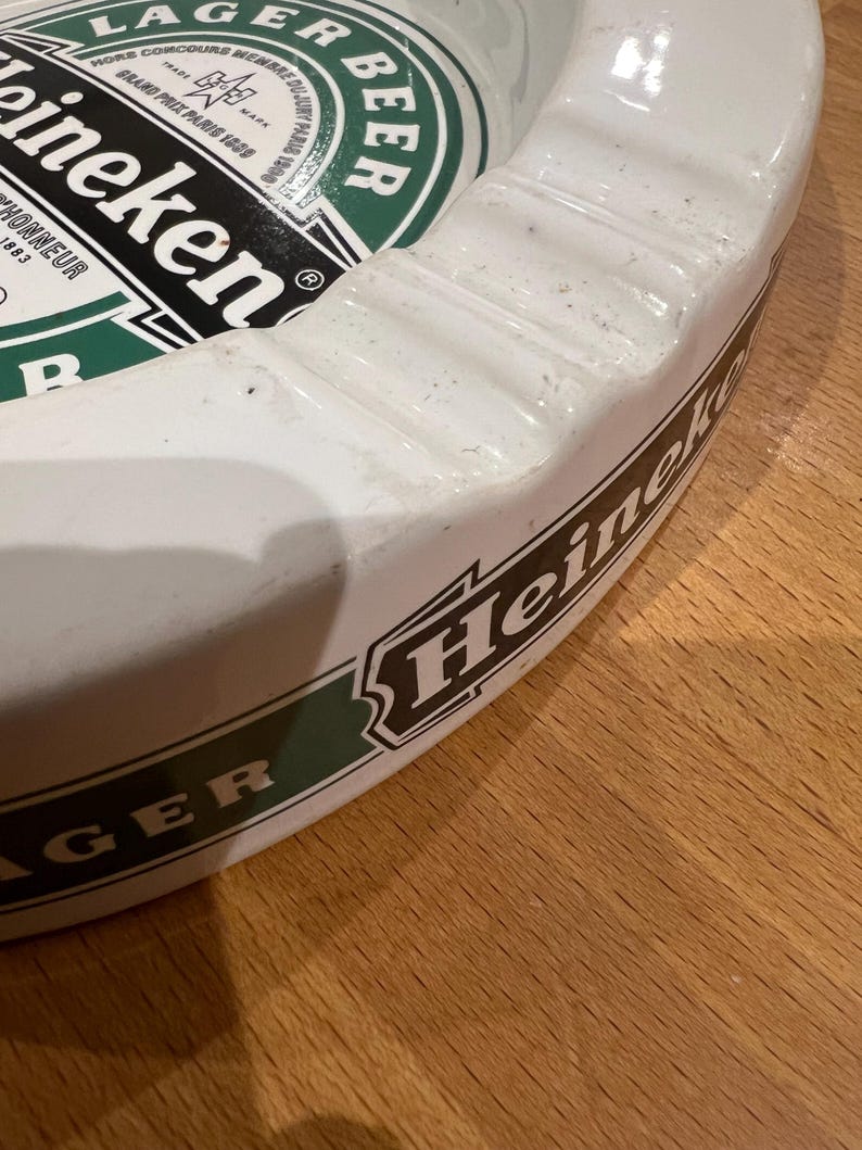 May include: White ceramic ashtray featuring the Heineken logo in green and black. The ashtray is circular with a raised edge and a grooved design. The words "LAGER BEER" and "Heineken" are displayed on the surface.