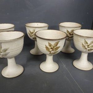 May include: Set of six white ceramic goblets with brown leaf designs. The goblets have a rounded bowl and a tall, slender stem.
