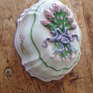 May include: A white ceramic mould shaped like a bunch of asparagus tied with a purple ribbon. The mould has a green and pink trim.