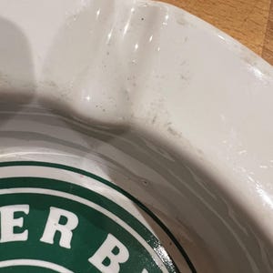 May include: A close-up shot of a white ceramic ashtray with a green and white logo. The ashtray has a curved rim and a circular design in the centre. The logo features the words "ER BE" in white letters.