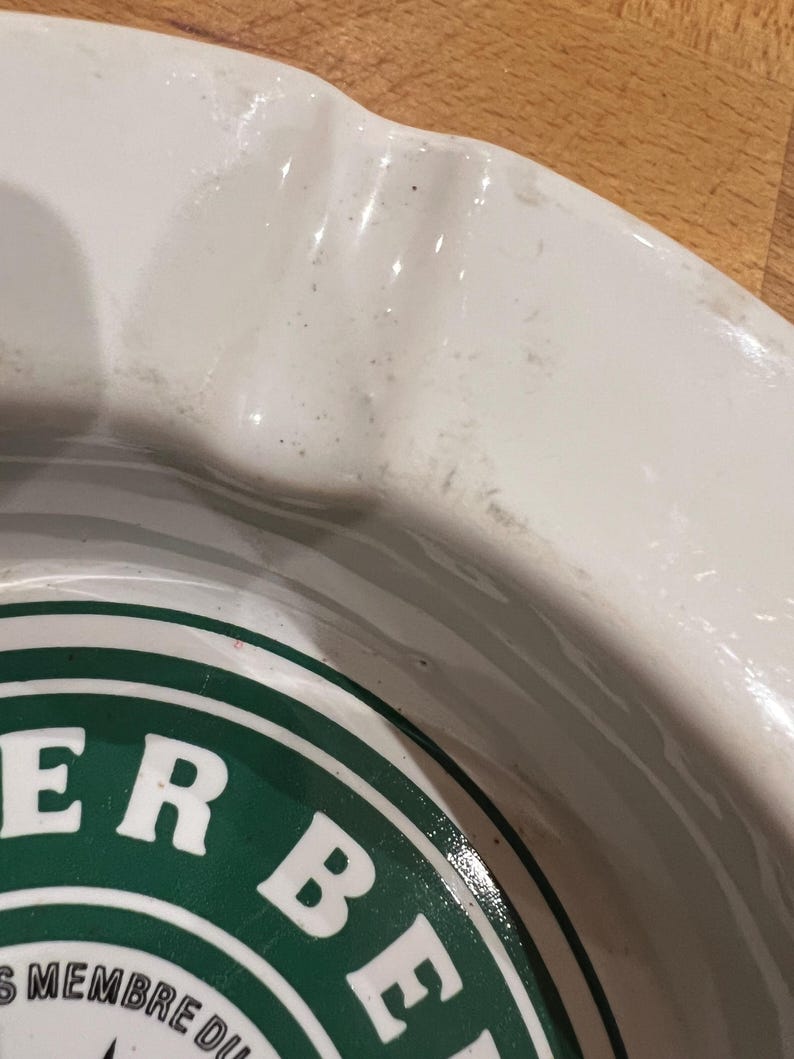 May include: A close-up shot of a white ceramic ashtray with a green and white logo. The ashtray has a curved rim and a circular design in the centre. The logo features the words "ER BE" in white letters.