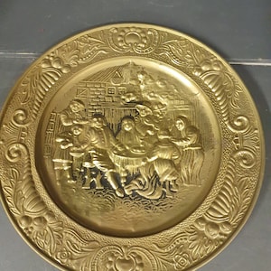 Könnte beinhalten: Eine goldfarbene Metallwandplatte mit einem geprägten Design von Menschen, die um einen Tisch versammelt sind. Die Platte hat einen dekorativen Rand mit floralen und Schriftrollenmotiven.