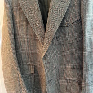 May include: A gray plaid blazer with a classic design. The jacket features a notched lapel, two front pockets, and a breast pocket. The fabric has a subtle pattern of red and white lines. The blazer is hanging on a wooden hanger.