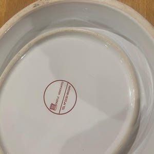 May include: A white ceramic serving dish with a distinctive, curved shape. The base displays a red circular logo with text. The dish has a glossy finish and a slightly aged look around the edges.
