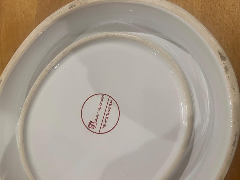 May include: A white ceramic serving dish with a distinctive, curved shape. The base displays a red circular logo with text. The dish has a glossy finish and a slightly aged look around the edges.