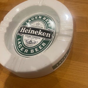 May include: White ceramic ashtray with a circular design. The centre features the Heineken logo in green and white, with the words "Lager Beer" around the edge. The ashtray has a curved shape with a lip for resting cigarettes.