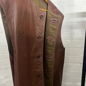 May include: A brown leather vest with a button-up front and a contrasting olive green lining. The vest is sleeveless and hangs on a wooden hanger. The leather has a worn appearance.