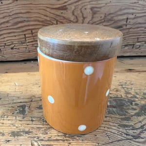 Może przedstawiać: Pomarańczowy ceramiczny słoik z białymi kropkami i drewnianą pokrywką. Słoik jest cylindryczny i stoi na drewnianej powierzchni. Pokrywka jest w jasnobrązowym kolorze.