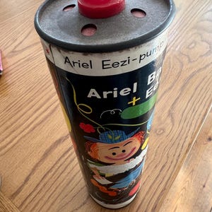 Puede incluir: Contenedor Ariel Eezi-pump vintage. El recipiente cil&iacute;ndrico tiene un fondo negro con ilustraciones y texto coloridos. La parte superior es de metal con una pieza de pl&aacute;stico roja. El texto en el contenedor dice "Ariel Eezi-pump".