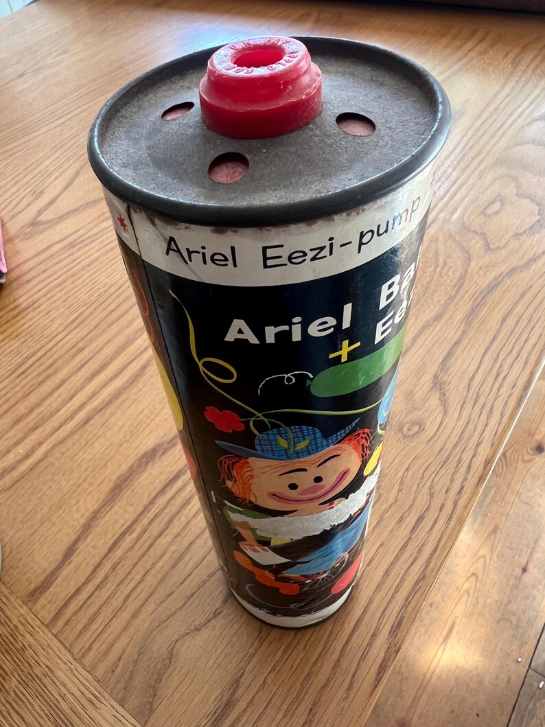 Puede incluir: Contenedor Ariel Eezi-pump vintage. El recipiente cil&iacute;ndrico tiene un fondo negro con ilustraciones y texto coloridos. La parte superior es de metal con una pieza de pl&aacute;stico roja. El texto en el contenedor dice "Ariel Eezi-pump".