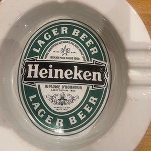 May include: White ceramic ashtray featuring the Heineken Lager Beer logo in green and black. The ashtray has a circular shape with a central depression and grooves for resting cigarettes. The logo includes text such as "Diplome D'Honneur Amsterdam 1883".