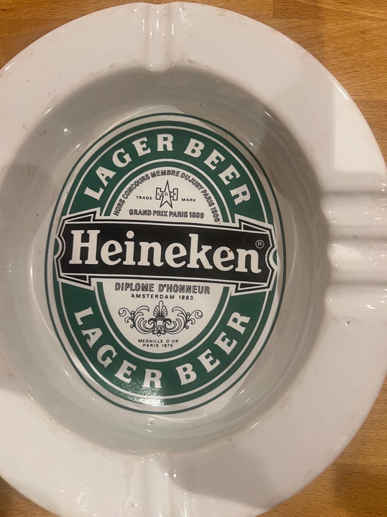 May include: White ceramic ashtray featuring the Heineken Lager Beer logo in green and black. The ashtray has a circular shape with a central depression and grooves for resting cigarettes. The logo includes text such as "Diplome D'Honneur Amsterdam 1883".
