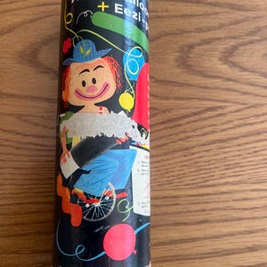 Puede incluir: Un recipiente cil&iacute;ndrico para un inflador de globos Ariel Eezi-pump. El recipiente es negro con una ilustraci&oacute;n de dibujos animados de un payaso y globos. El texto "Ariel Balloon + Eezi-pump" est&aacute; impreso en el recipiente.