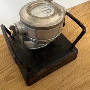 May include: A vintage fuel level meter in silver and black, with a black metal stand. The meter has a circular face with a needle and a dial that reads "Fuel Level Meter". The stand has a rectangular base and two vertical supports.