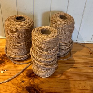 May include: Three spools of natural-coloured rope are displayed on a wooden surface. The rope is tightly wound around cardboard cores. The spools vary in size, with one spool unwound, extending a length of rope across the wood.
