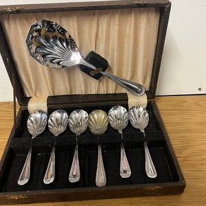 Può includere: Un set di sette cucchiaini d'argento con bordi a festoni e un cucchiaio da portata in argento abbinato, il tutto in una scatola di legno marrone con fodera beige.