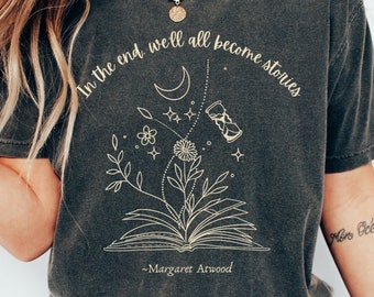 May include: A dark gray t-shirt with a graphic design of an open book with a flower growing out of it. The graphic also includes a crescent moon, stars, and an hourglass. The text on the shirt reads "In the end, we'll all become stories - Margaret Atwood".