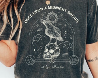 May include: A dark gray t-shirt with a graphic print of a raven perched on a skull. The graphic is surrounded by stars, clouds, and a crescent moon. The text "Once upon a midnight dreary - Edgar Allan Poe" is printed below the graphic.