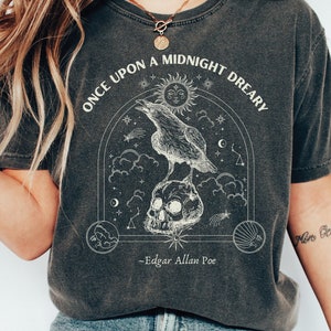 May include: A dark gray t-shirt with a graphic print of a raven perched on a skull. The graphic is surrounded by stars, clouds, and a crescent moon. The text "Once upon a midnight dreary - Edgar Allan Poe" is printed below the graphic.