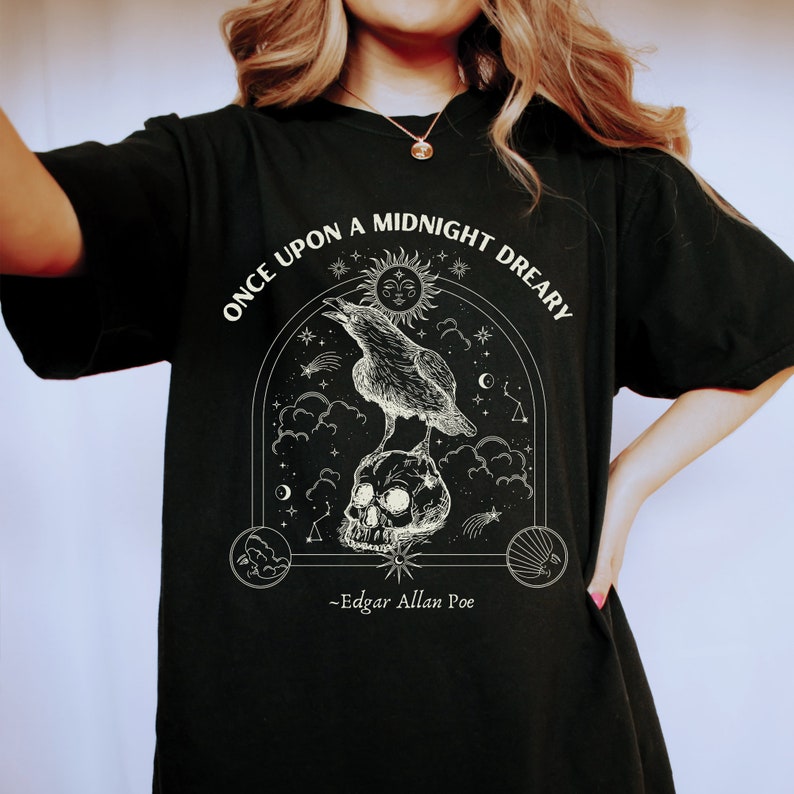 May include: Black t-shirt with a white graphic design featuring a raven perched on a skull with a crescent moon and stars. The text reads "Once upon a midnight dreary" and "Edgar Allan Poe".