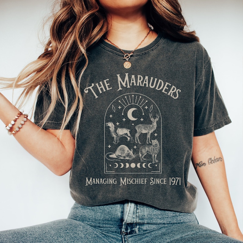 May include: A dark gray t-shirt with a vintage-style graphic featuring the text "The Marauders" and an illustration of four animals: a dog, a deer, a rat, and a wolf. The graphic also includes a crescent moon, stars, and the text "Managing Mischief Since 1971".